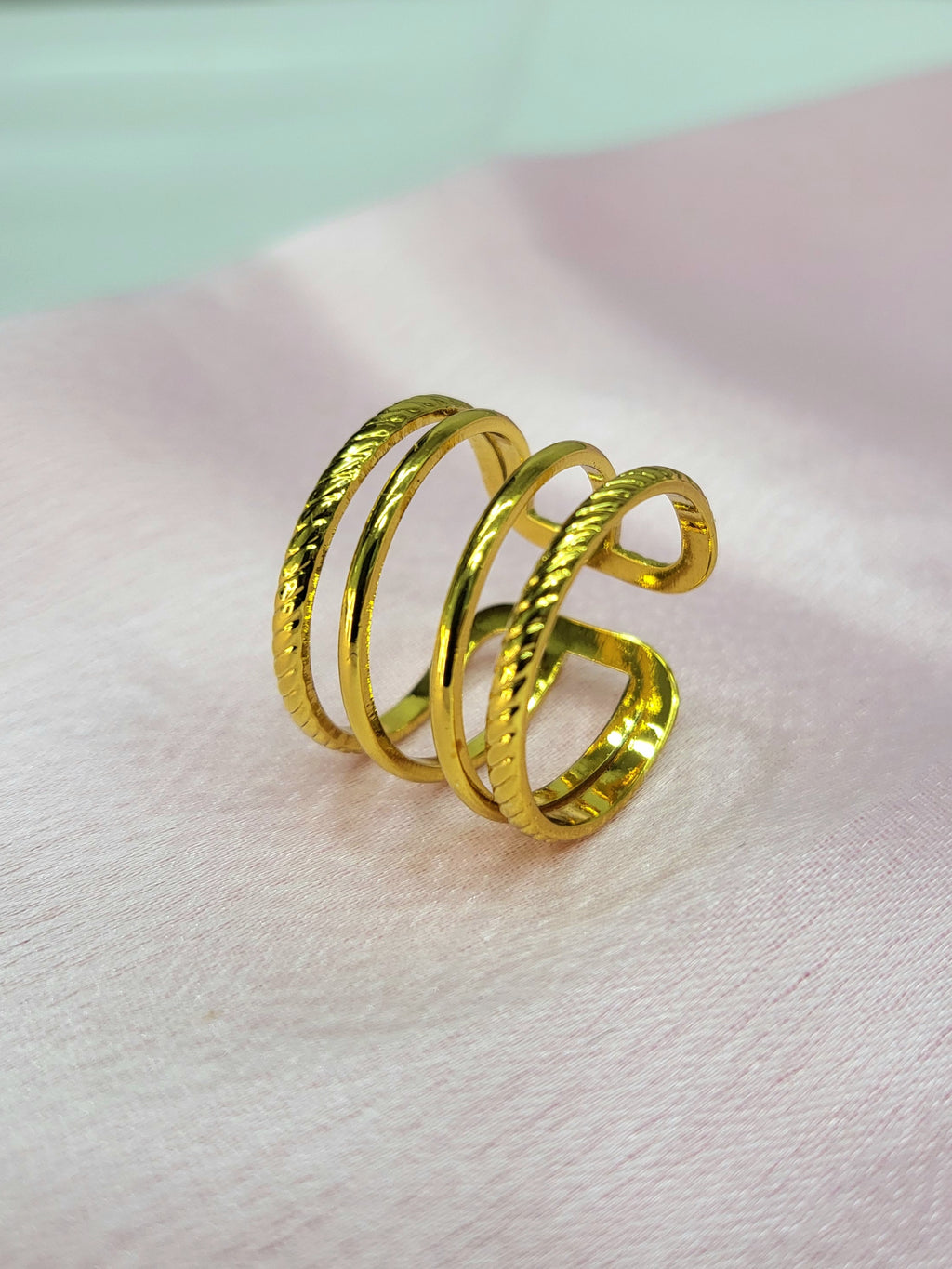 Lyvoria adjustable anti-tarnish gold plated stainless steel extra-wide cage ring featuring a four-row design with alternating twisted rope and smooth wires on a soft pink background.