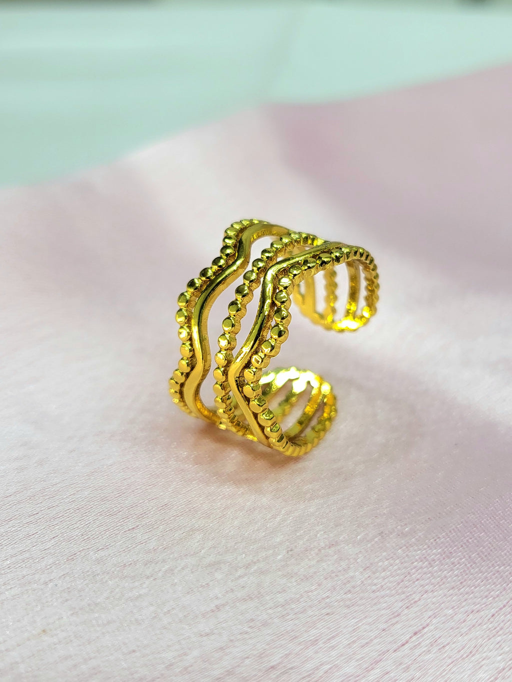 Estelle adjustable anti-tarnish gold plated stainless steel wide cage ring featuring a multi-row wave design with alternating smooth and beaded gold rows on a soft pink background.